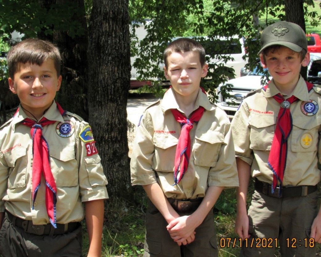 Three scouts in tan uniforms with red neckerchiefs standing outdoors near trees.