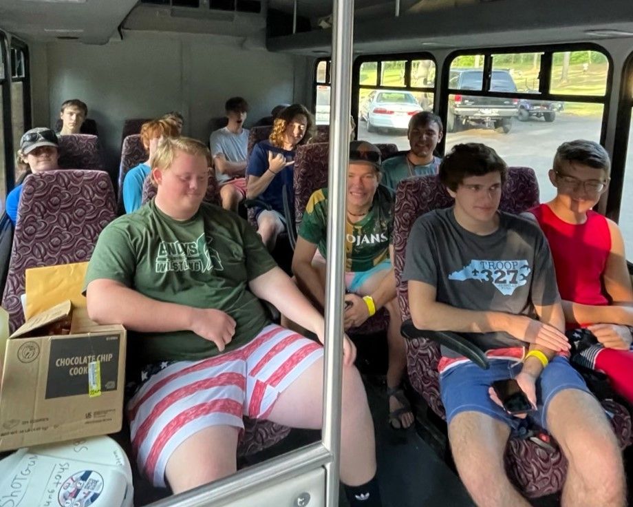 Passengers seated on a bus, some smiling and chatting, with backpacks and a cardboard box nearby