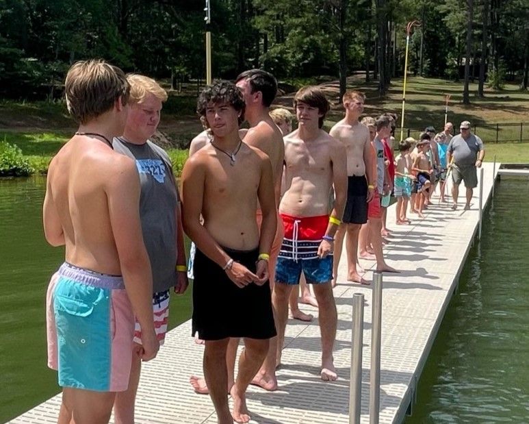 People standing in a line on a dock beside a lake, wearing swim trunks and towels.