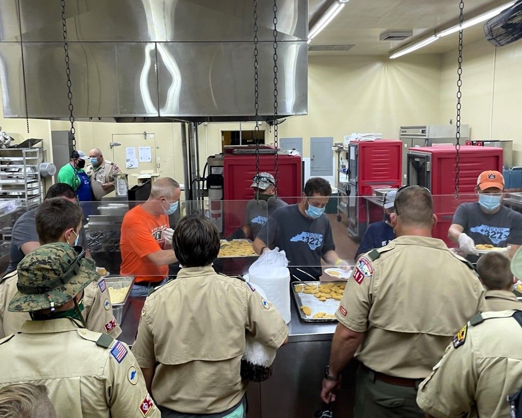 People in uniforms and masks serving food at a buffet counter in a kitchen or cafeteria