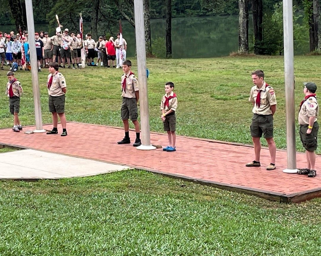 Scouts standing on a red brick path in a grassy outdoor camp area, with others gathered in the background.