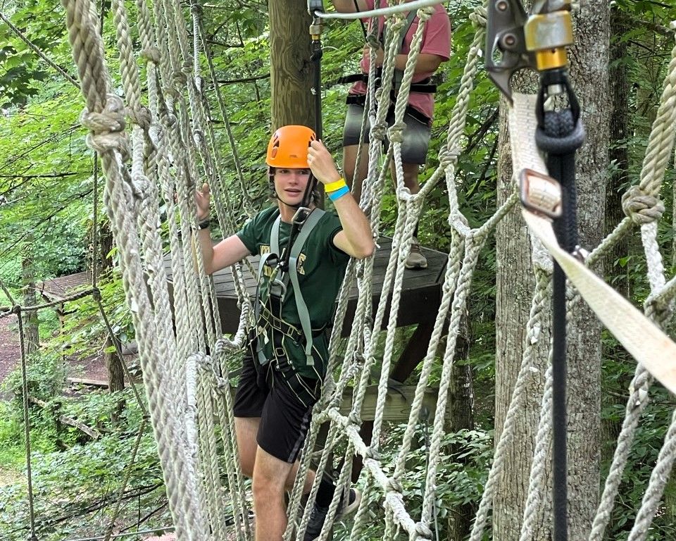 Person in orange helmet crossing a rope bridge in a forest obstacle course