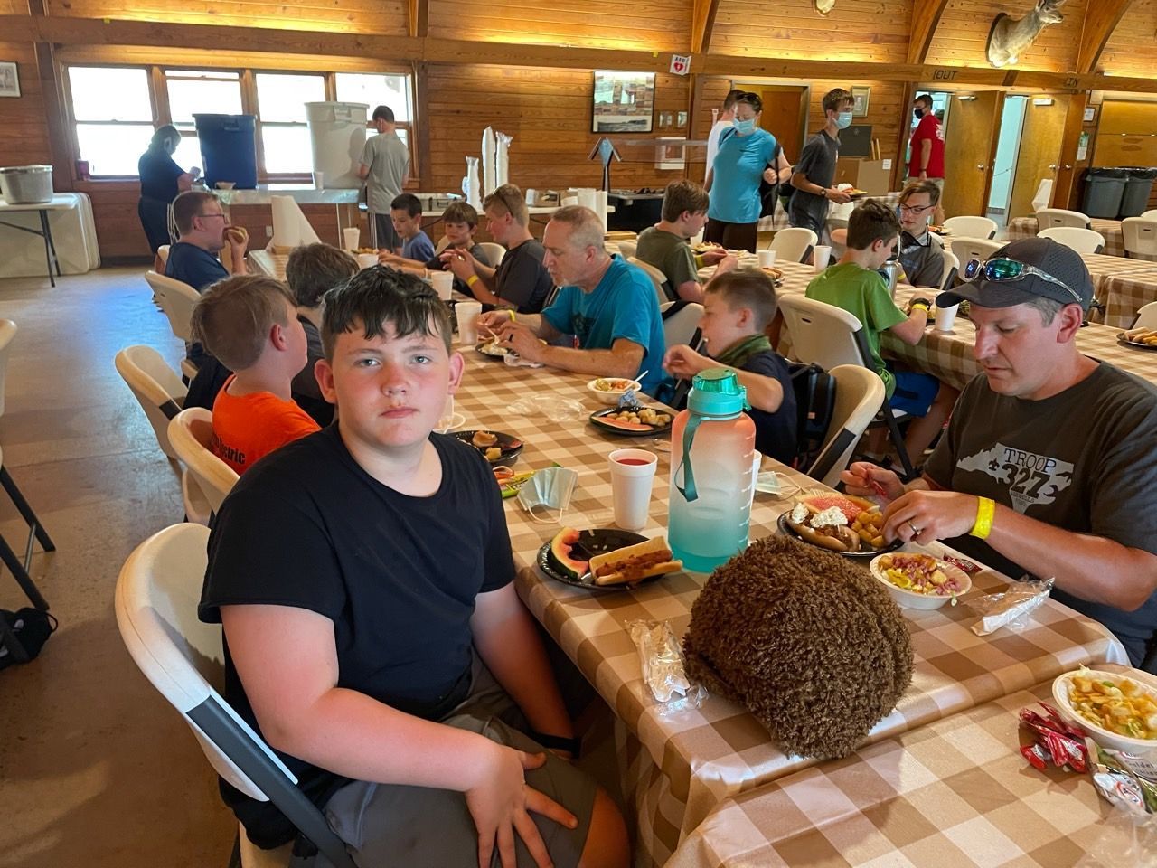 People seated at a long table in a rustic dining hall, sharing a meal with trays and drinks.