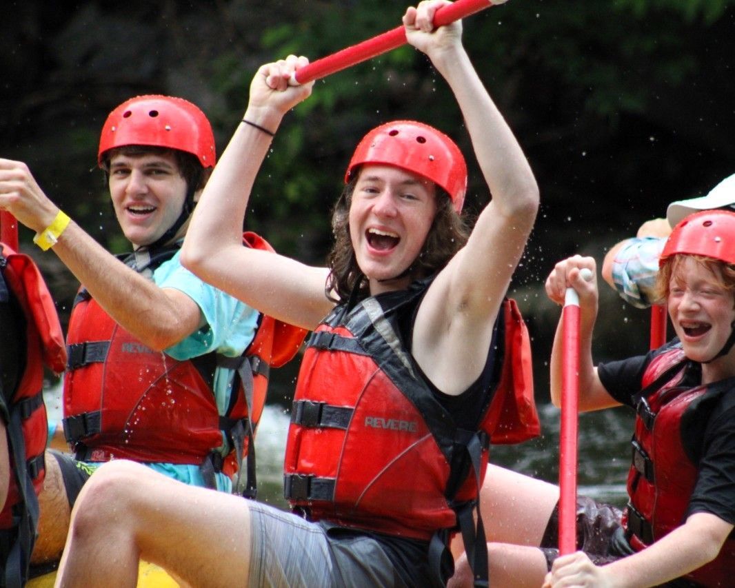 Three people in red helmets and life vests cheer while paddling a raft on whitewater.