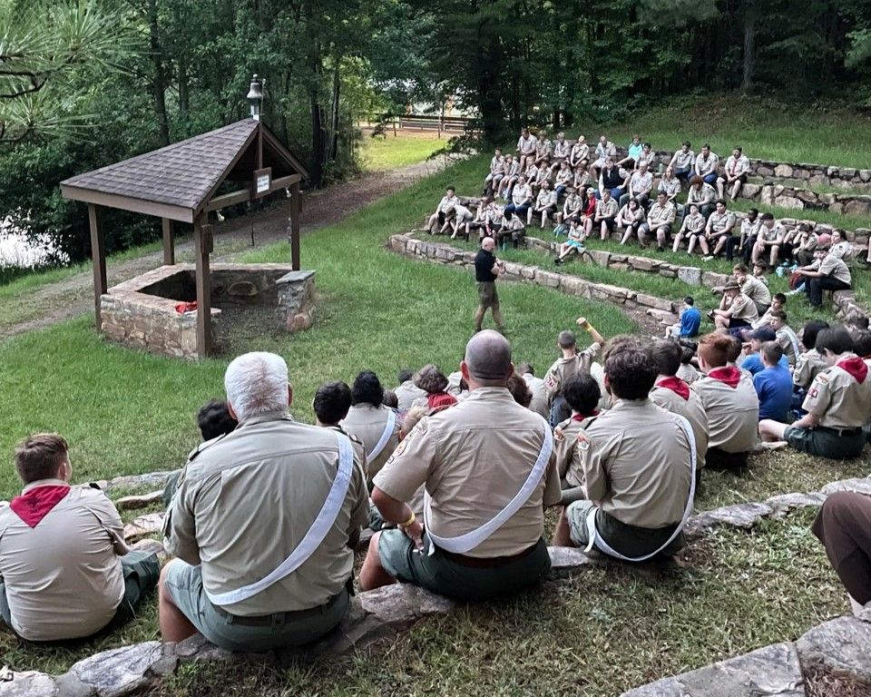 Outdoor group gathering in a wooded amphitheater, with people seated on a grassy slope facing a speaker.