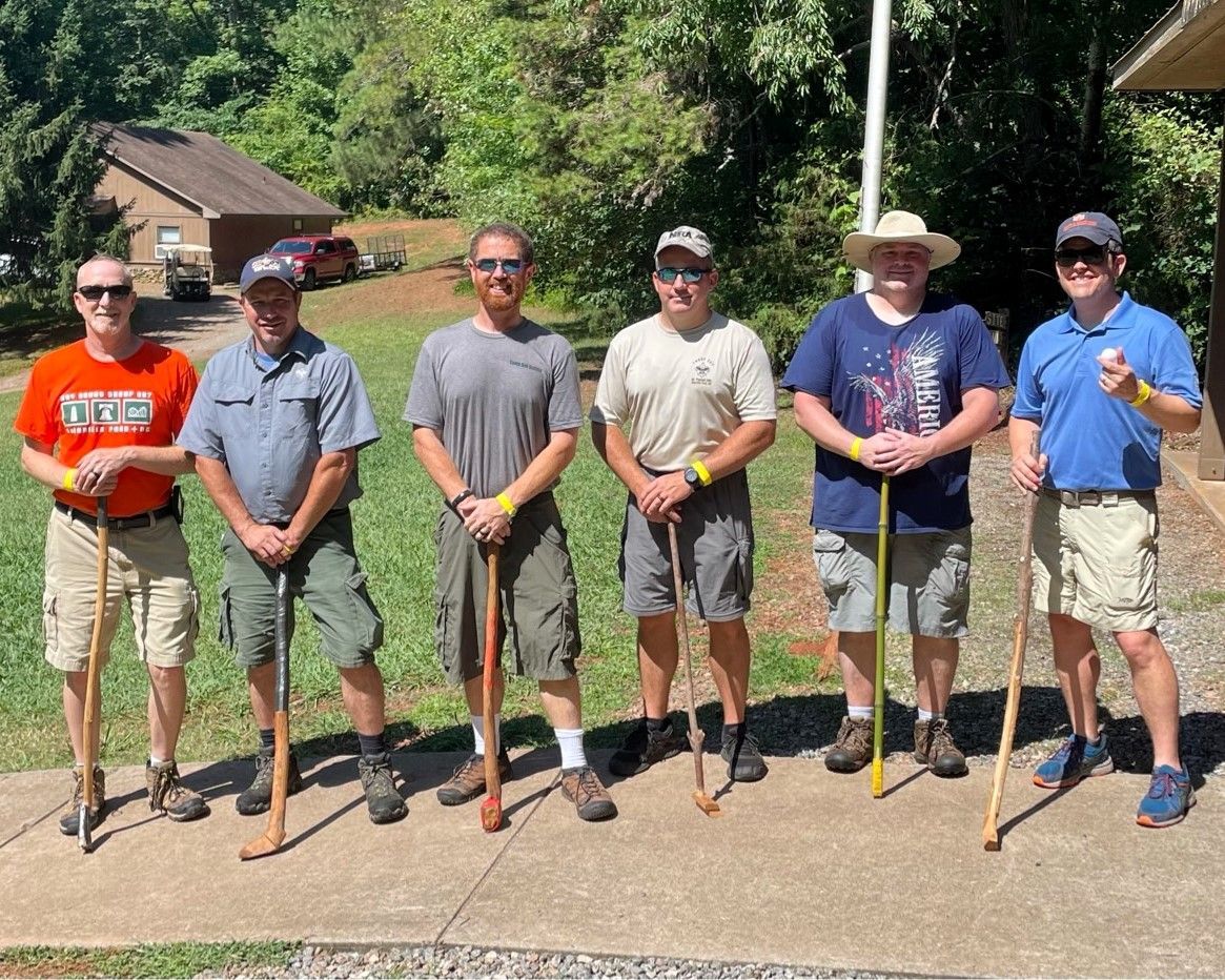 Six people stand outdoors on a sunny lawn, posing with shovels in front of a house.