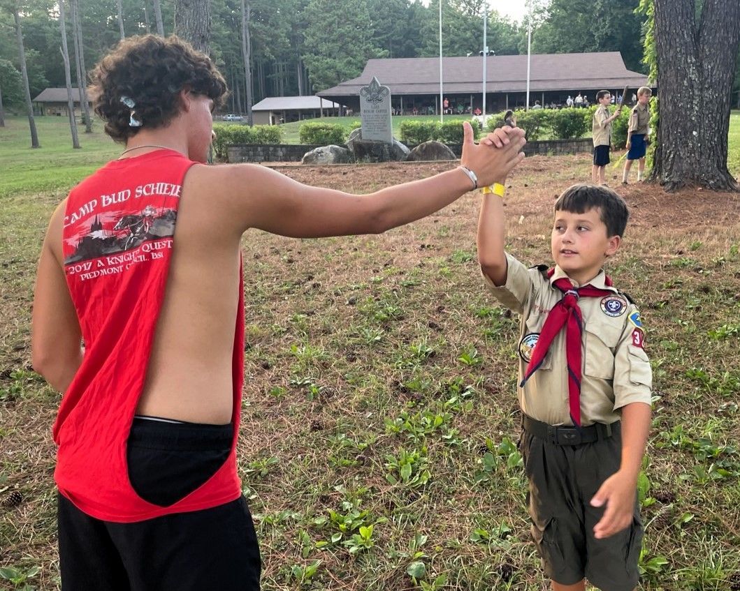 Two scouts in uniform high-five outdoors near trees and a building