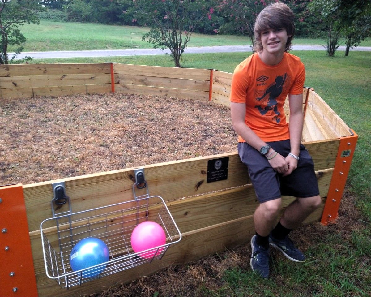 Boy sitting beside a wooden fenced enclosure with two colorful balls in a grassy park.