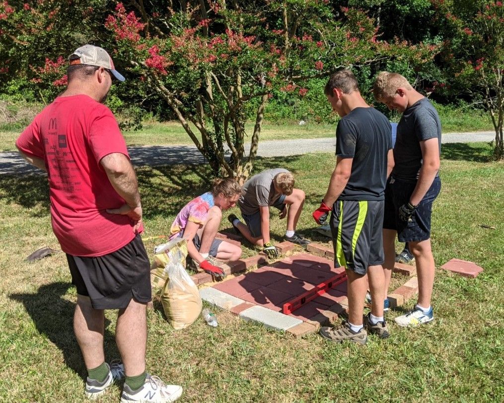 People kneel around a red-and-white ground marker in a sunny park while others watch nearby