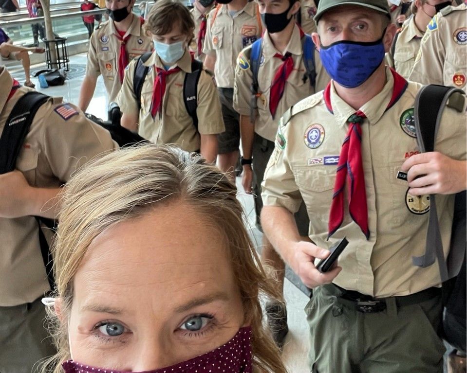 Masked Scouts in tan uniforms and red neckerchiefs standing together in a bright indoor hallway