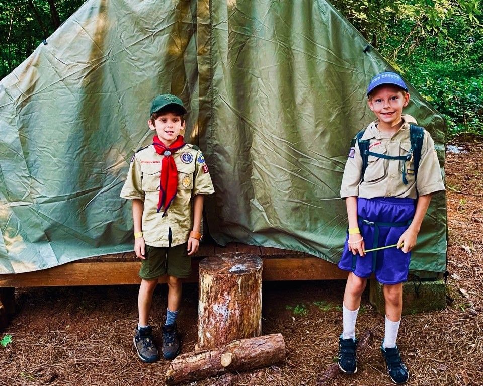 Two scouts in khaki uniforms stand in front of a green tarp shelter in the woods.