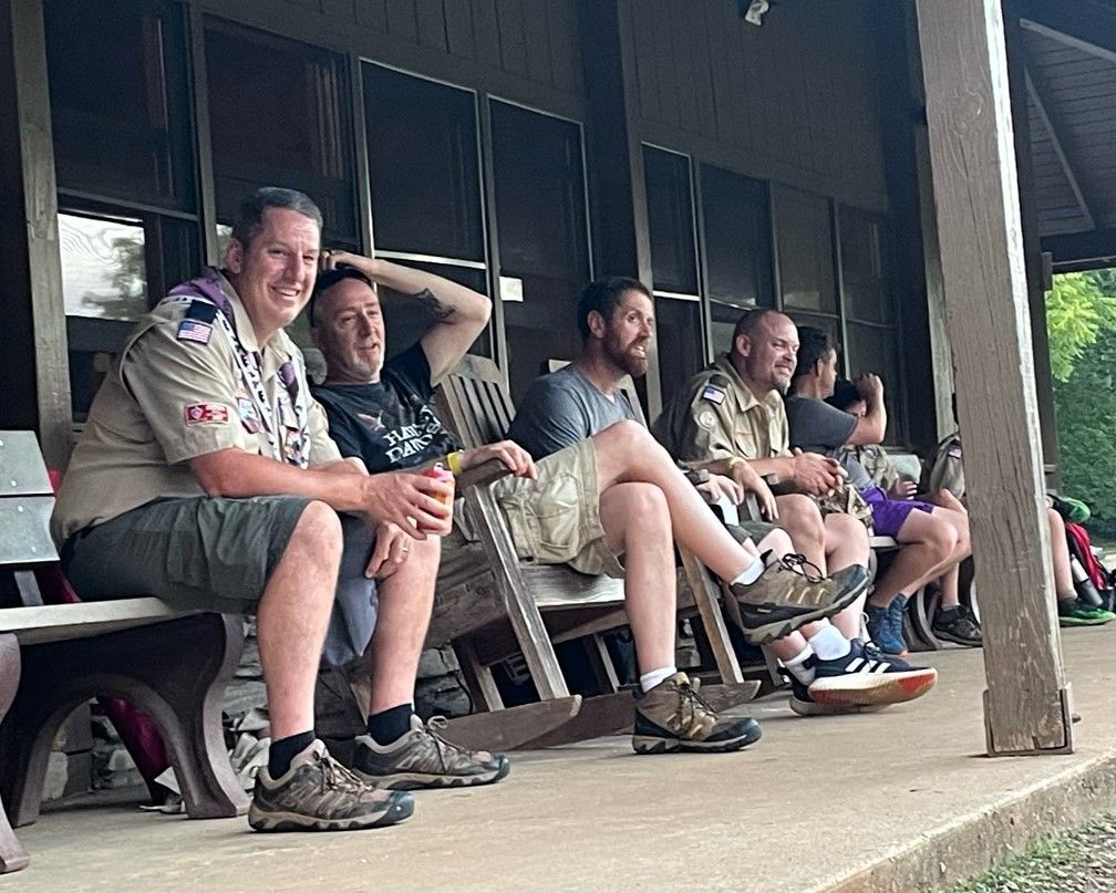 Several people in scout uniforms sit on a porch, resting and chatting.