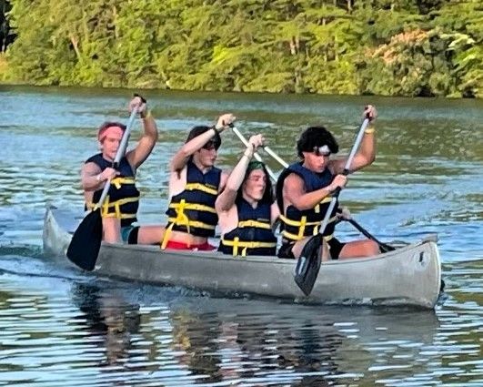 Four people in life jackets paddling a canoe on a calm lake with trees in the background