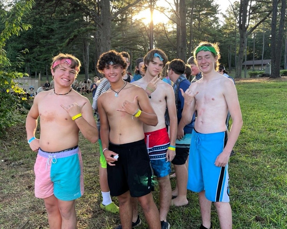 Five shirtless friends posing outdoors at sunset, wearing colorful swim trunks and wristbands.