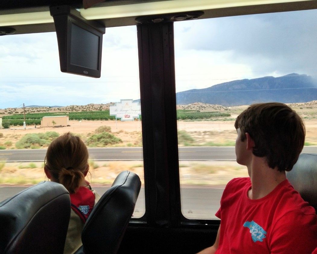 Two children in red shirts ride a bus, looking out at a desert landscape and mountains.