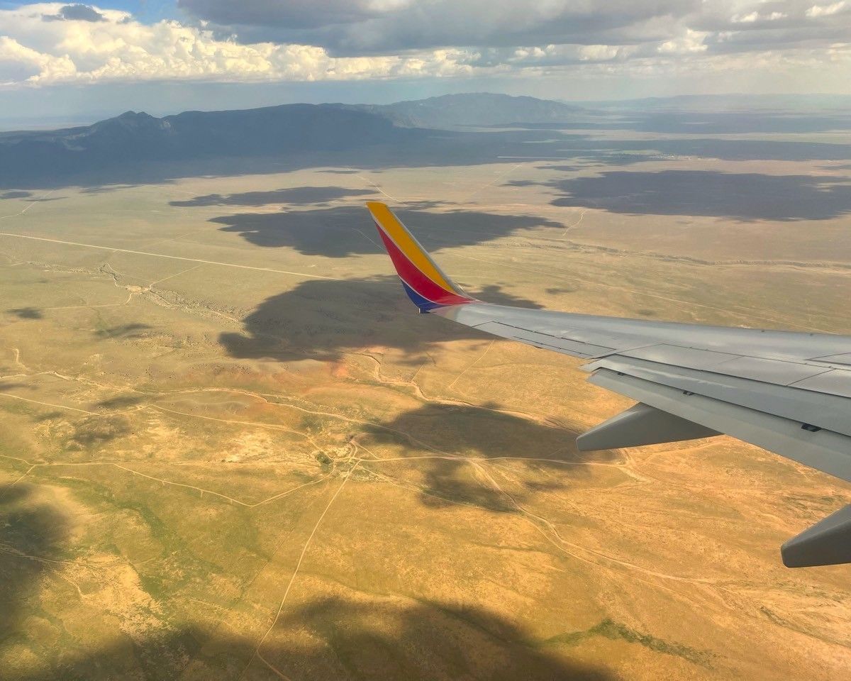 Airplane wing over a sunlit desert landscape with distant mountains and clouds