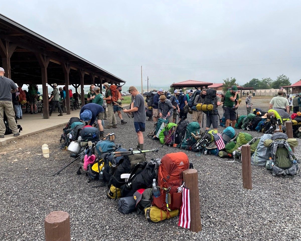 Backpacks lined up on gravel beside a covered platform as people gather in a transit area