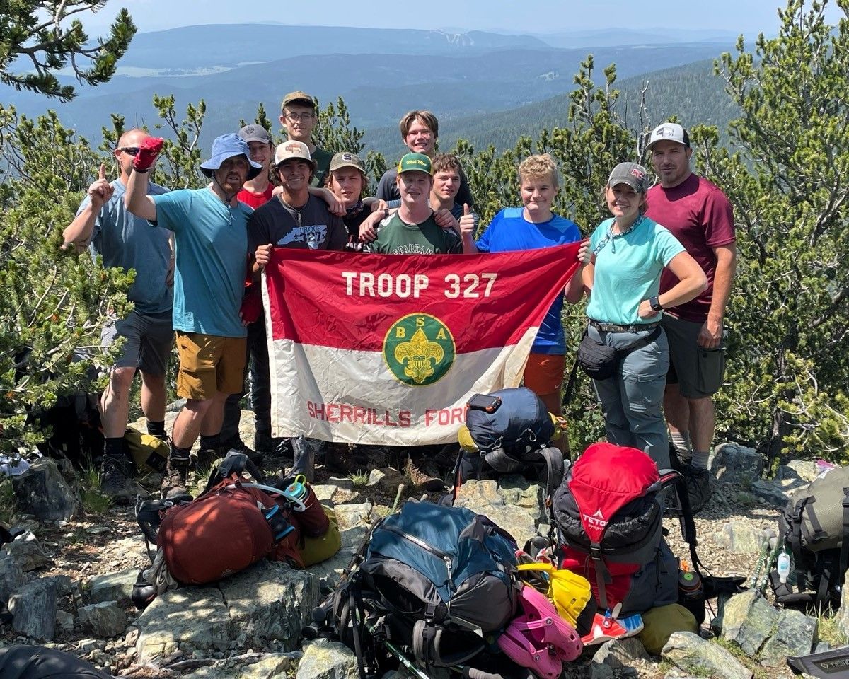 Hikers posing on a mountaintop with Troop 327 banner and backpacks, overlooking distant hills.