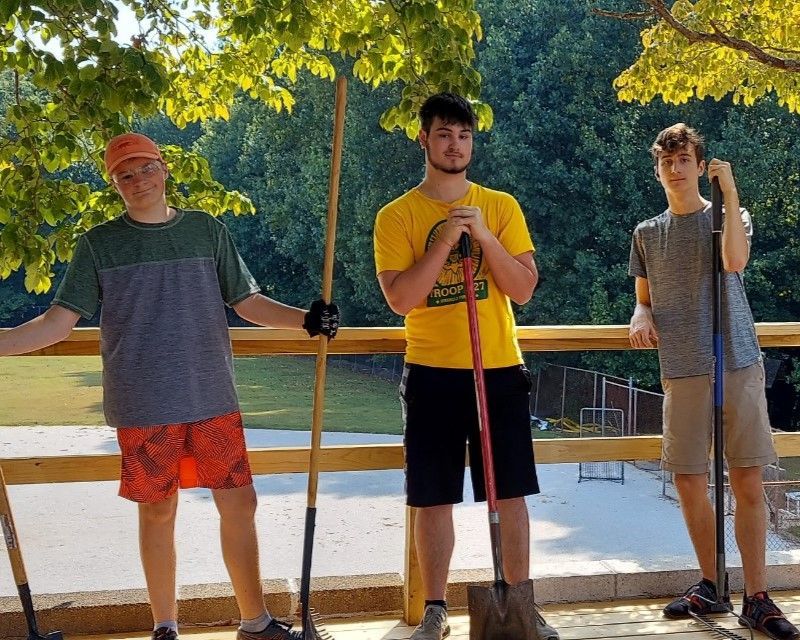 Three people holding paddles stand on a dock beside a lake under trees.