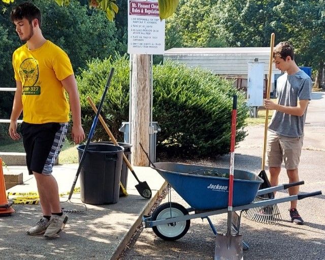 Two people work outdoors near a blue wheelbarrow, shovels, and buckets on a gravel path.