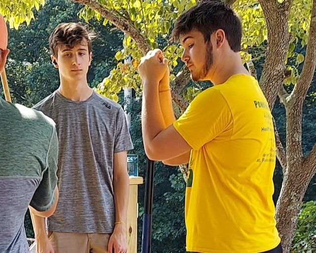 Two people outdoors, one in a yellow shirt gesturing while talking to another in a gray shirt.