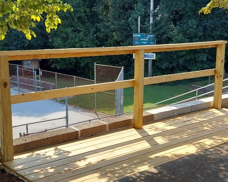 Wooden deck with railing overlooking a fenced tennis court and trees beyond.