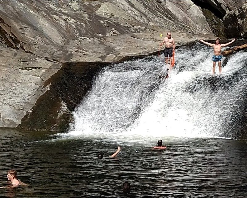 People swimming and standing near a small waterfall in a rocky pool.