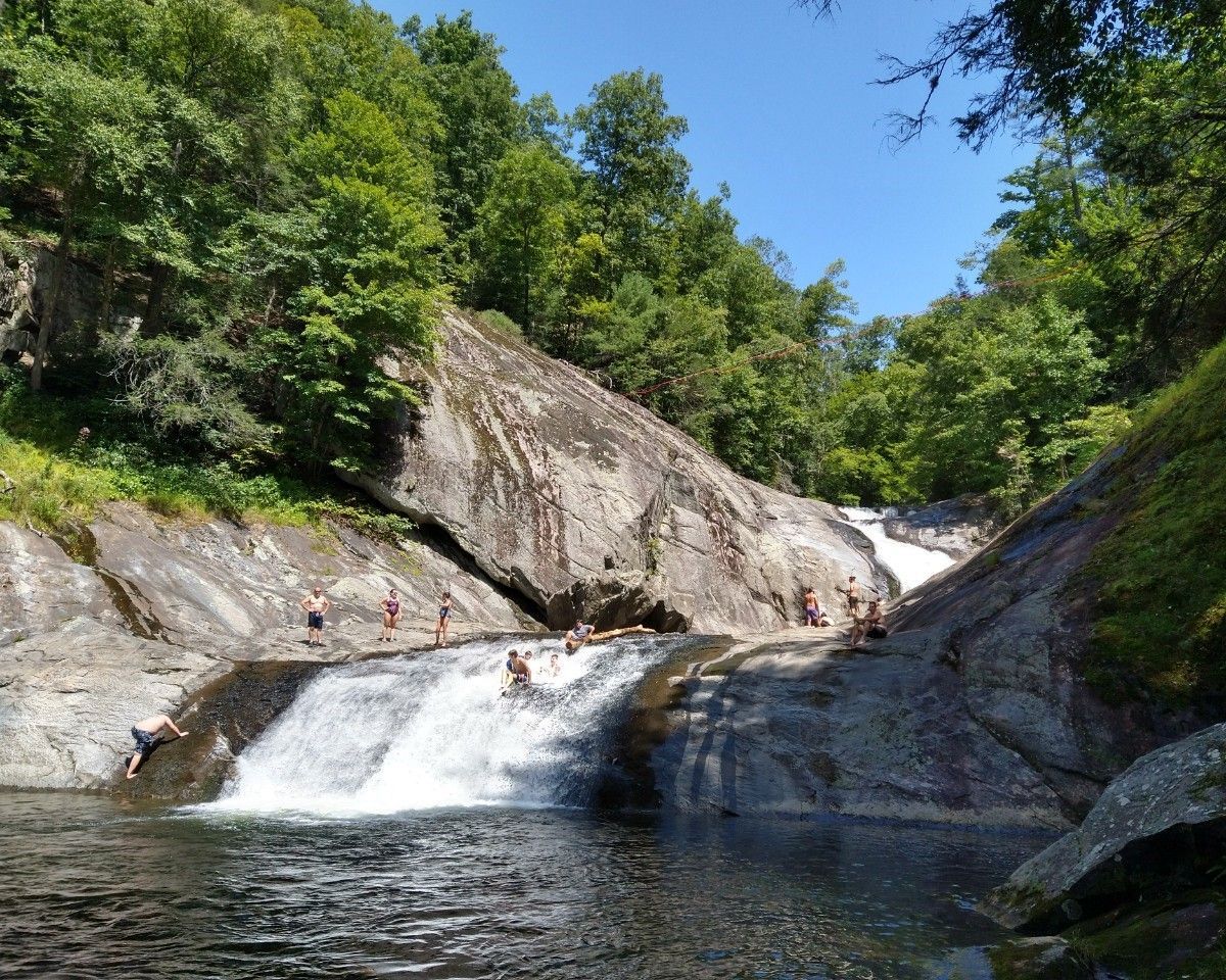 Small waterfall flowing over rock ledges into a clear pool, surrounded by green forest under a blue sky