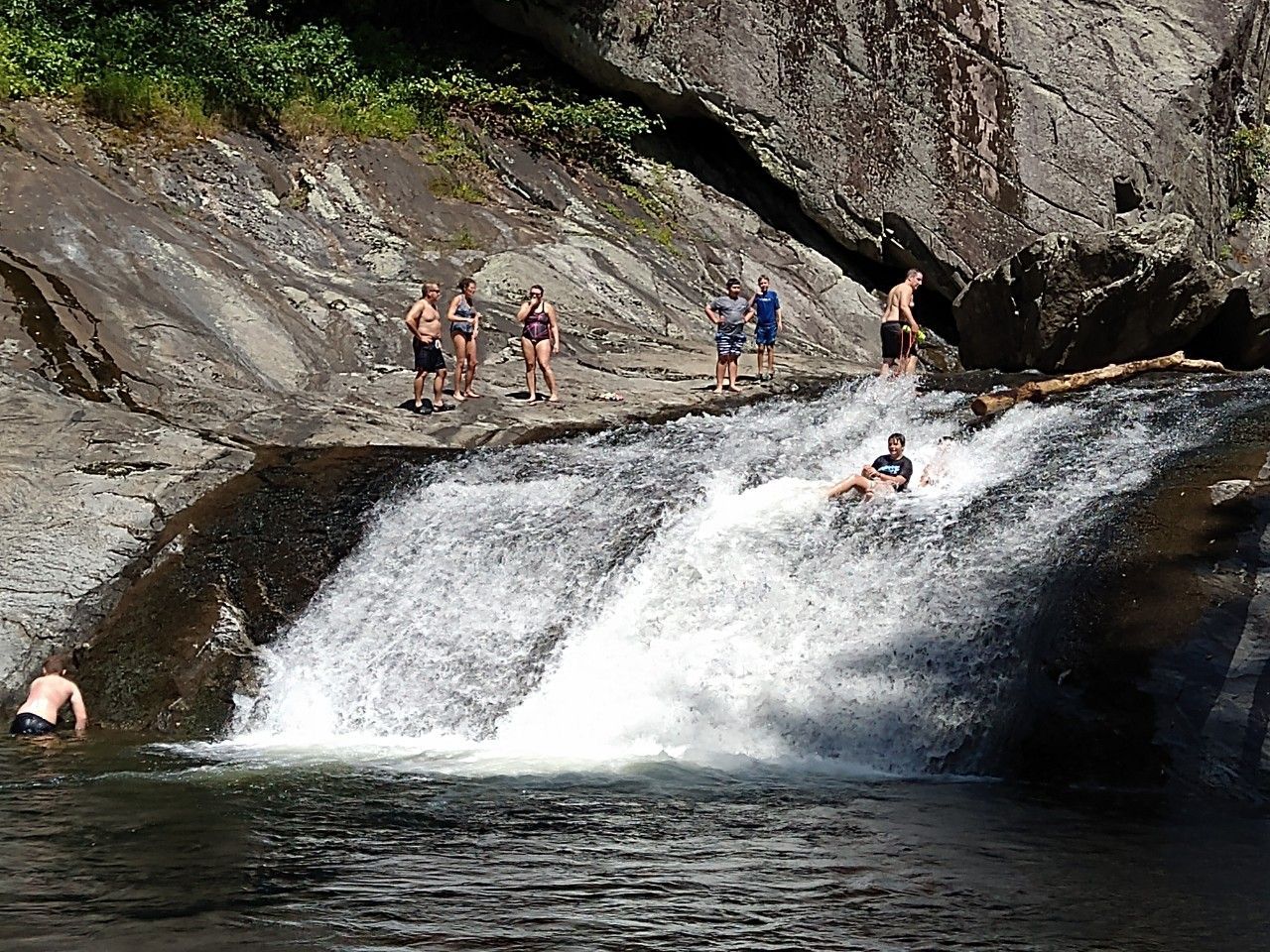 People swimming and standing on a rocky waterfall ledge above a pool