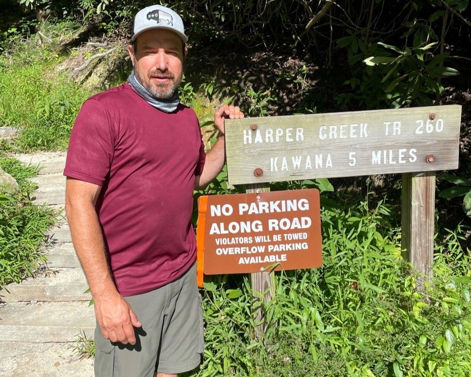 Man standing beside trail signs reading “Harper Creek Tr 260” and “No parking along road”
