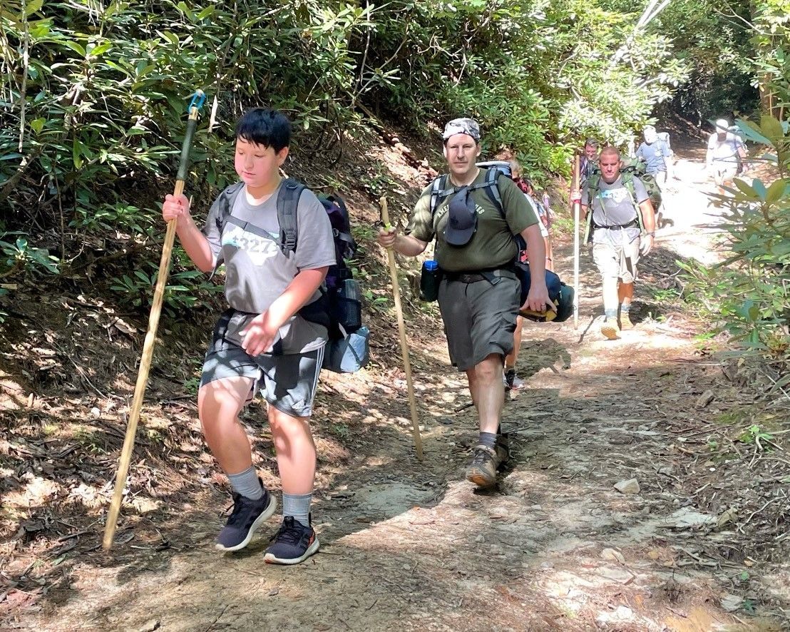 Hikers with backpacks and walking sticks hiking along a wooded trail in bright sunlight