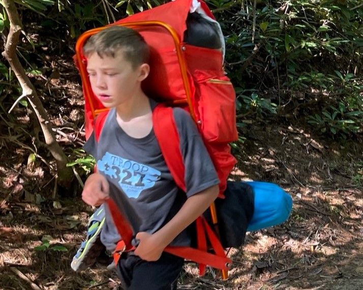 Child hiking on a wooded trail with a large red backpack and a blue sleeping roll