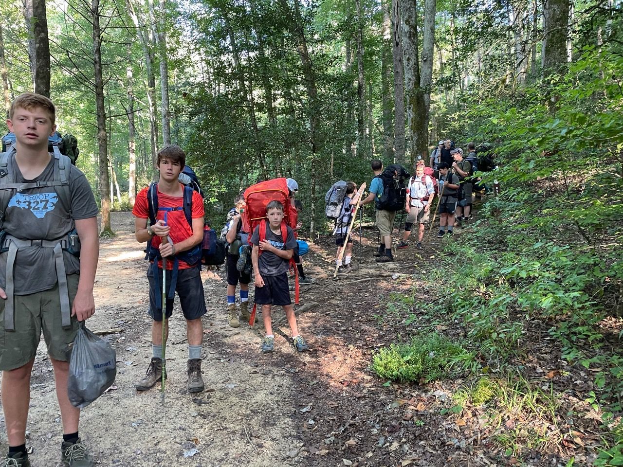 Hikers with backpacks walking along a forest trail, some bending over and resting in the woods.