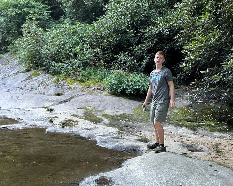 Person standing on a rocky stream bank beside water and dense green trees