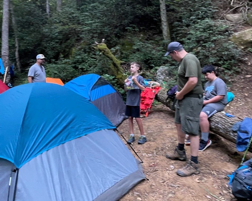 Campers gather beside tents in a wooded campsite, with backpacks and gear on the ground.