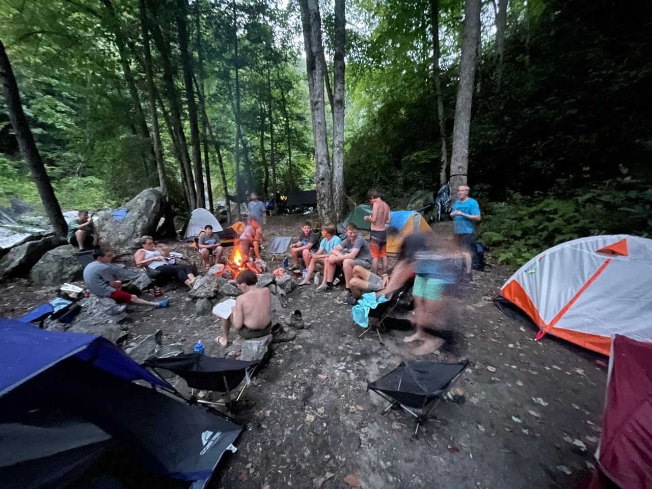 Campers gathered around a campfire in a wooded campsite, with tents and rocks nearby.