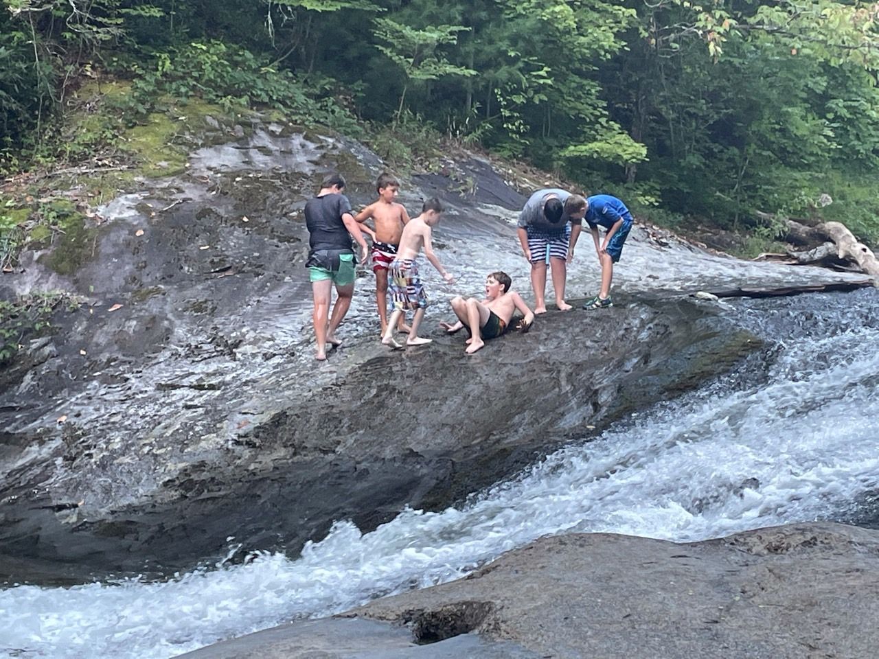 People standing and crouching on a rocky waterfall ledge by a forest stream.