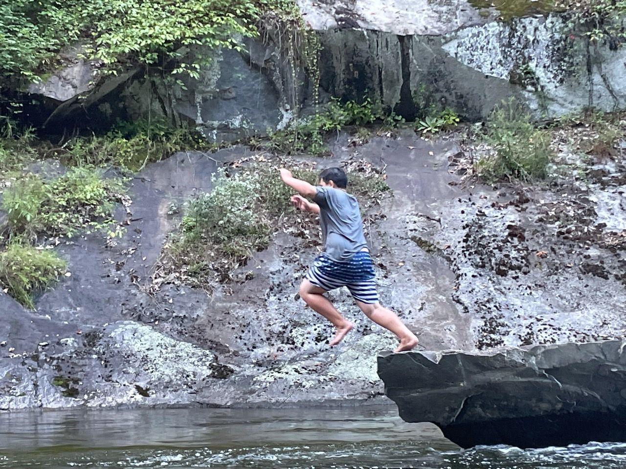 Person jumping from a rock ledge over water near a rocky, green riverbank