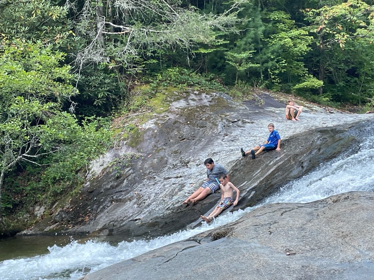 People sliding down a rocky stream chute into white water, surrounded by green forest.