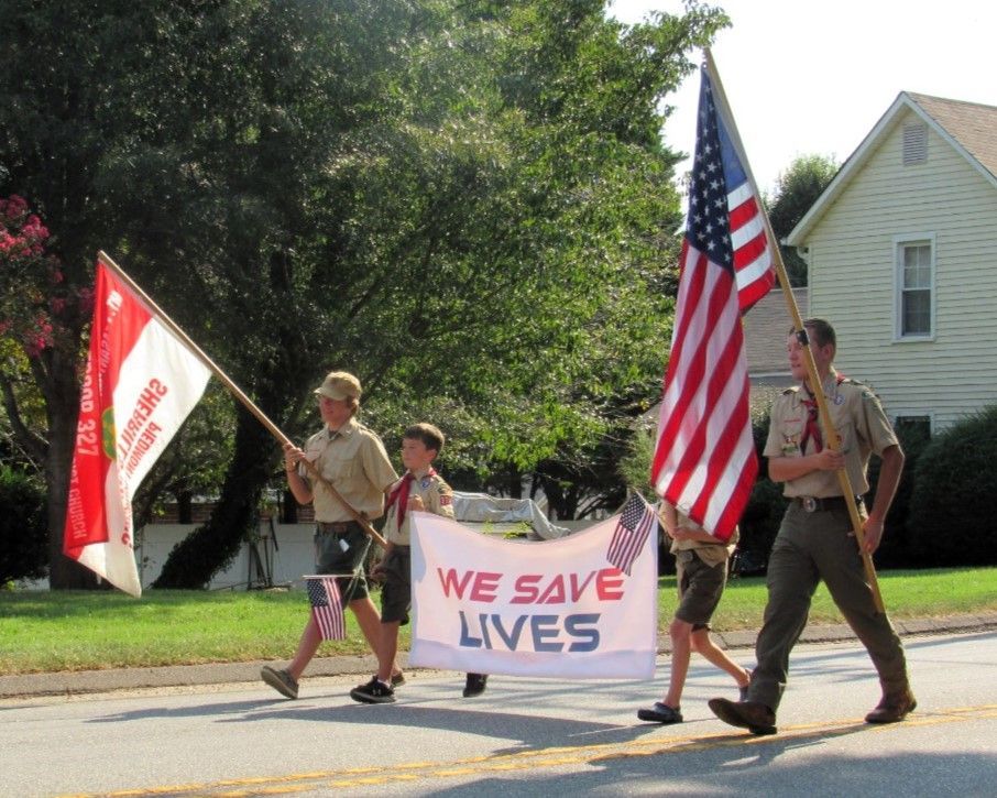 People marching with flags and a “WE SAVE LIVES” banner on a sunny street.