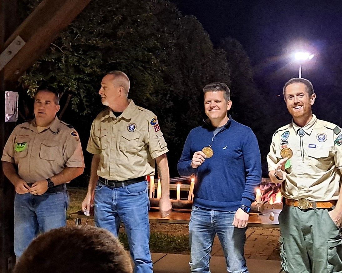 Four scouts in uniforms stand outdoors at night, one holding a microphone under bright lights.