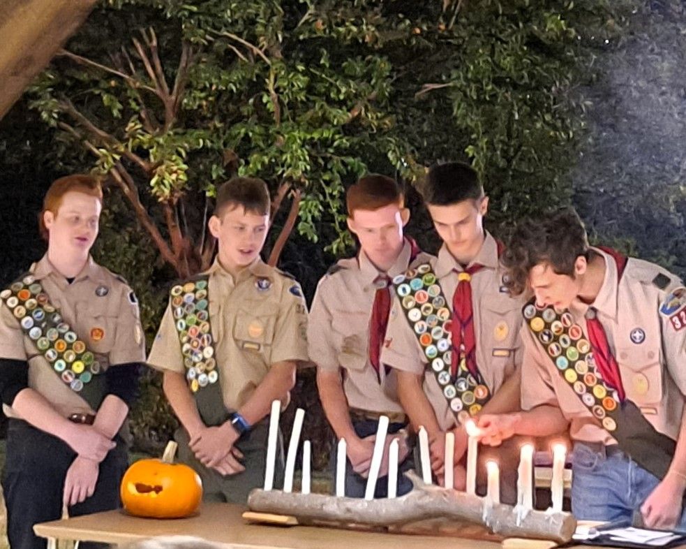 Five Scouts in tan uniforms sitting by lit candles and a pumpkin at a nighttime outdoor ceremony