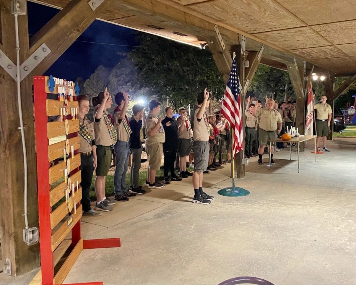 Uniformed scouts standing at attention in a covered pavilion during a flag ceremony, with an American flag visible.