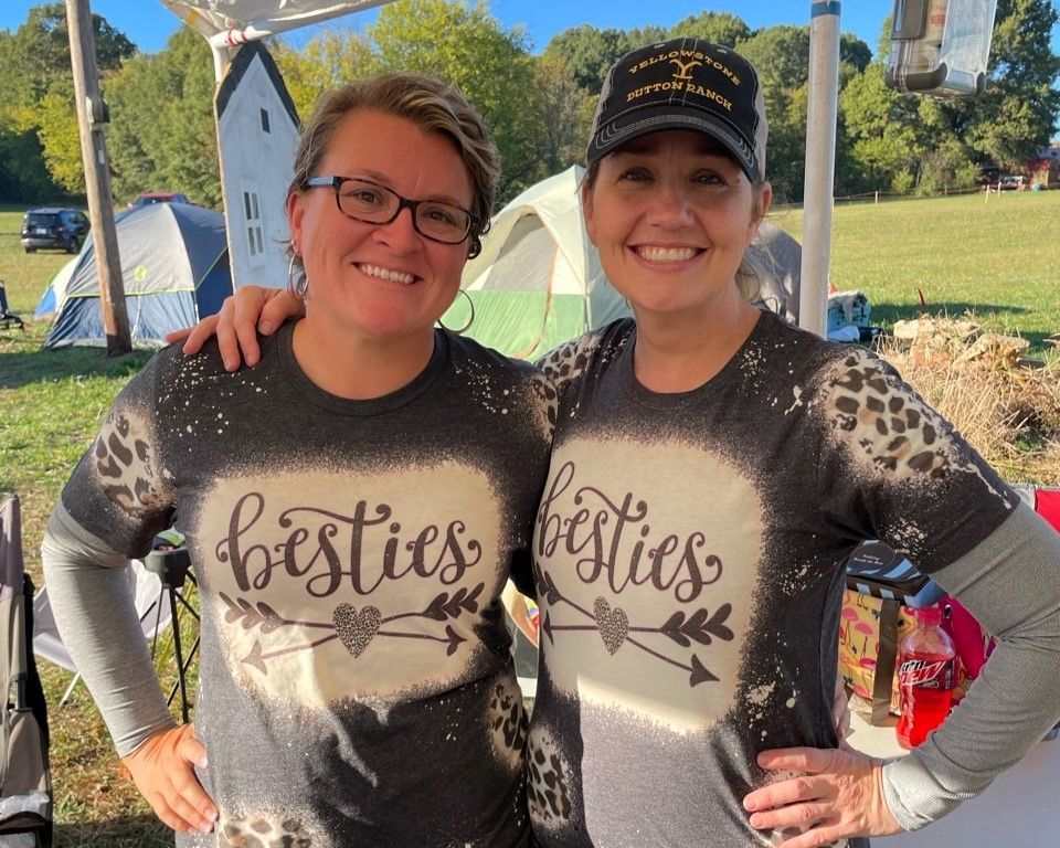 Two smiling people in matching “besties” shirts posing outdoors at a festival
