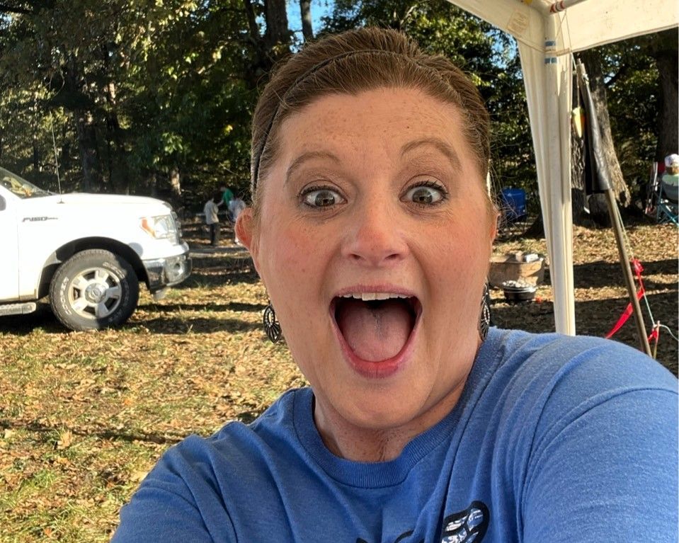 Smiling person in a blue shirt outdoors under a canopy, with a white truck in the background.