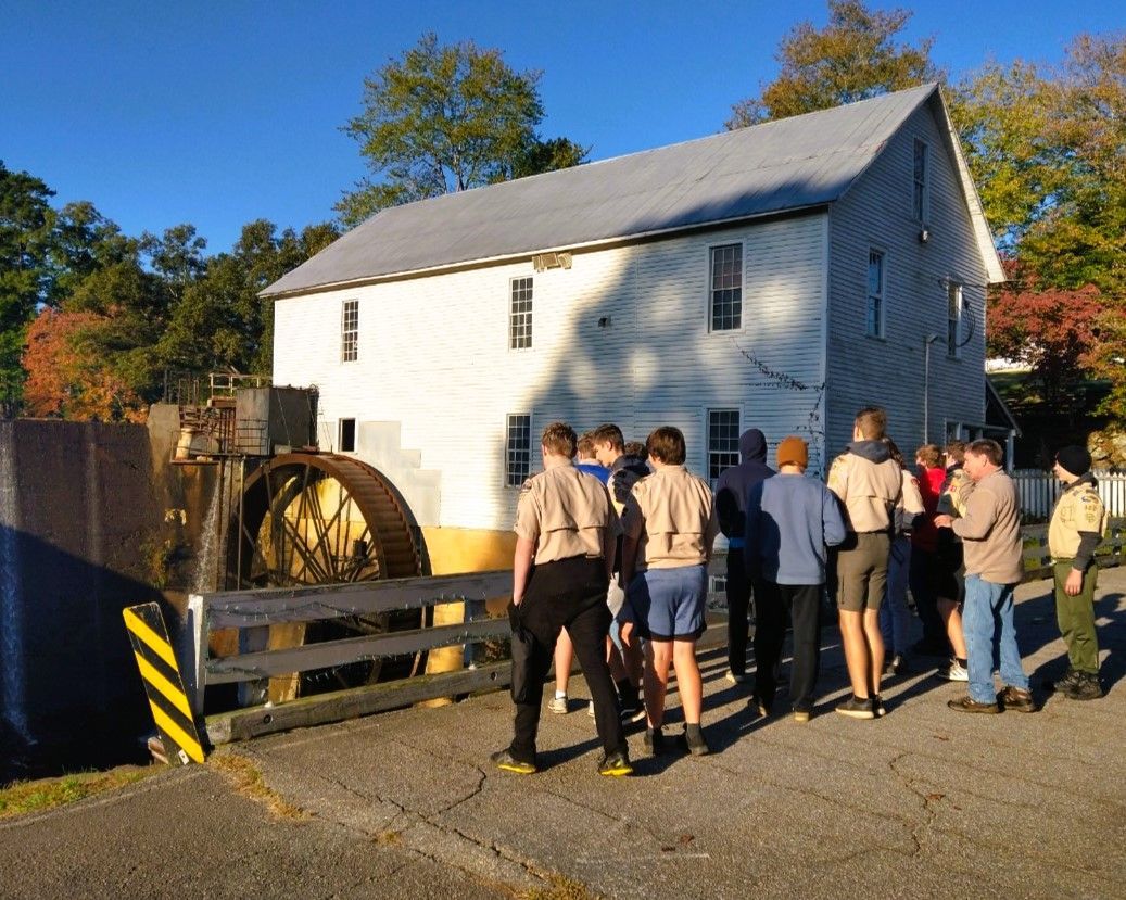 Group of people touring a white mill building beside a large water wheel on a sunny day