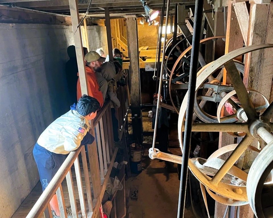 People standing on a narrow platform beside large industrial machinery and wheels in a dim workspace