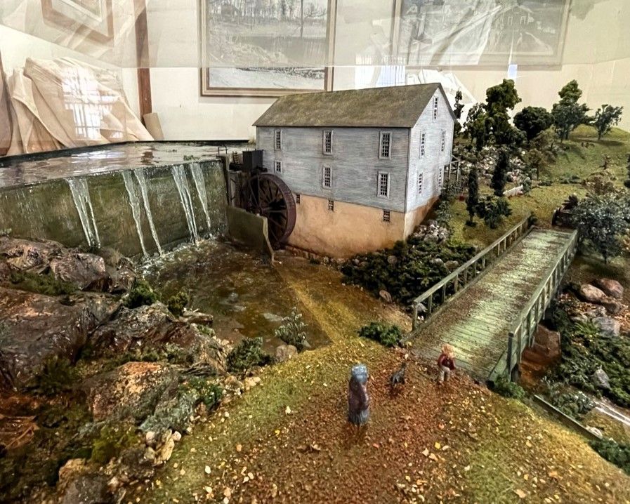 Miniature diorama of a rural stone mill with waterfall, stream, bridge, and tiny figures on a path