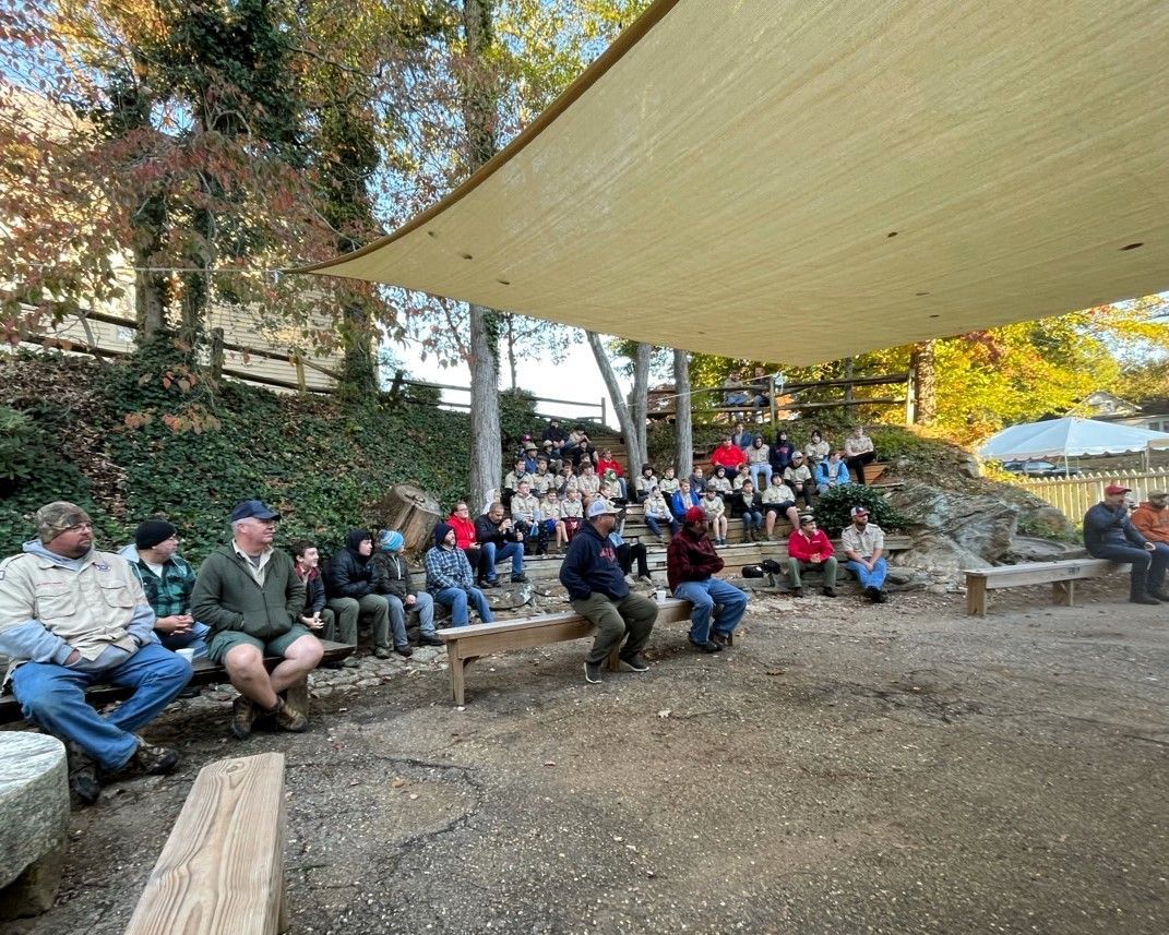People seated on benches outdoors under a large beige shade sail in a wooded park area.