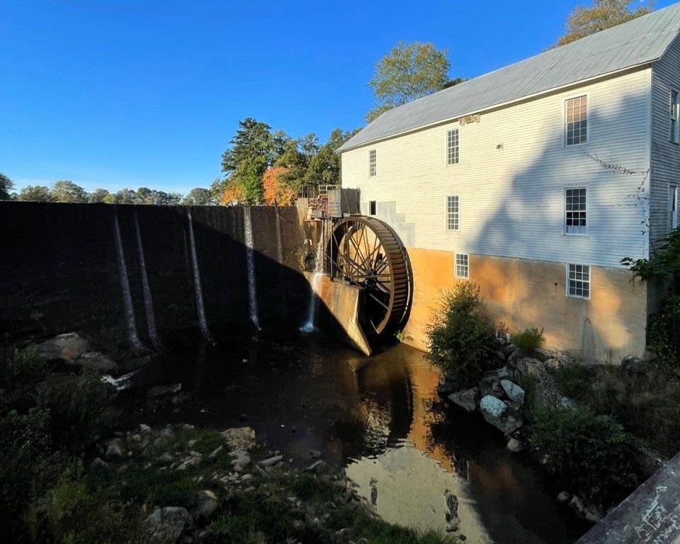 Waterwheel beside a white mill building over a canal, with a large dark wall casting shadow on the water.